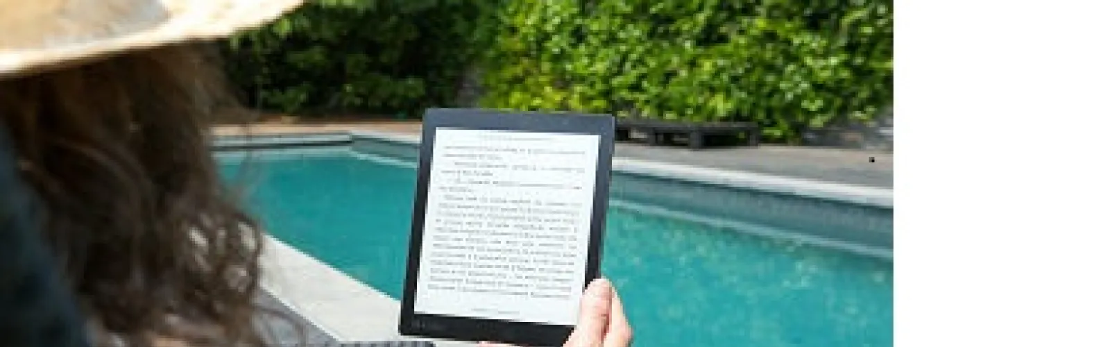 woman wearing hat by pool with e-reader